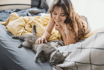 Attractive young woman plays and pets a British shorthair gray cat while lying in bed. Pet and care