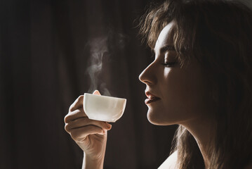 Pretty young woman holding a cup of espresso coffee from which steam is coming out on a dark background and enjoying the aroma