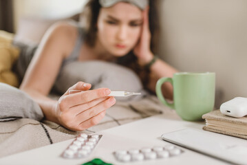 A young woman lies in bed and blows her nose into a handkerchief, takes her temperature and reaches for the pills with her hand. Self-isolation and sick leave at home. Cold