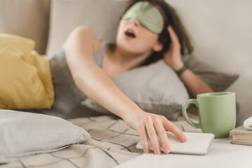 Fototapeta na wymiar Yawning young woman in a sleep mask reaches for her smartphone while lying in bed in a sunny bedroom. Turn off the alarm on your smartphone