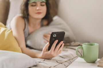 Sleepy young woman in a sleep mask reaches for her smartphone while lying in bed in a sunny bedroom. Turn off the alarm on your smartphone