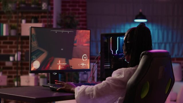 Over shoulder view of african american gamer girl playing online games using gaming pc setup spending relaxing time at home. Woman using modern technology for enterteinment and streaming tournament.