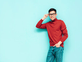 Fototapeta premium Portrait of handsome smiling model. Sexy stylish man dressed in red turtleneck sweater and jeans. Fashion hipster male posing near blue wall in studio. Isolated