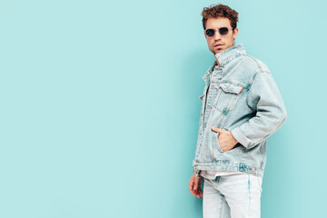 Portrait of handsome confident stylish hipster lambersexual model with curly hairstyle. Sexy man dressed in jeans jacket. Fashion male isolated on blue wall in studio. In sunglasses