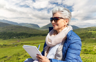 travel, tourism and vacation concept - happy senior woman writing to notebook over Killarney National Park valley in ireland background