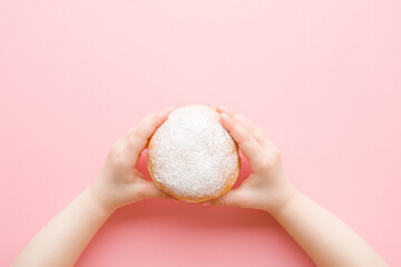 Baby girl hands holding fresh donut with white sprinkled sugar on light pink table background....