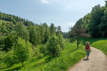 Woman with brown hair, straw hat,  gray t-shirt and jeans, hiking with red backpack on a trail to Staatspark Fürstenlager, rear view, Auerbach, Bensheim, Germany