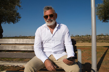 Handsome mature man with beard and grey hair is sitting thoughtfully on a park bench while consulting his mobile phone. The man is dressed in beige trousers, white shirt. Concept of old age.