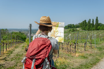 Woman with brown hair, gray t-shirt, jeans, straw hat and red backpack looking at a hiking map,...