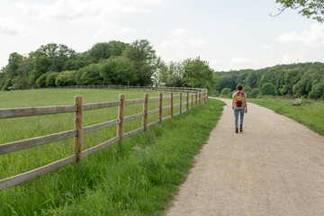 Obraz premium Woman with brown hair, gray t-shirt, jeans and straw hat attached to a red backpack hiking next to a paddock in the nature, rear view