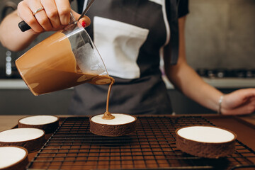 Smiling female confectioner pouring liquid chocolate gommage on biscuit cake. Woman chef in uniform at assembling mousse dessert process
