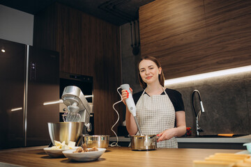 Young woman uses mixer and whips cream for cake, stands in modern kitchen