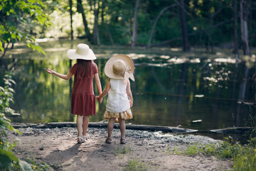 Children of a girl in hats and dresses in the summer look at the forest lake, back view. Summer walks in the parks