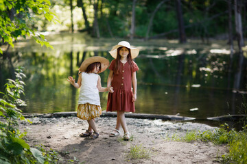Fototapeta premium Children girls in hats and dresses in summer together near forest lake. Summer calm walks in parks and forest