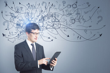 Portrait of attractive thoughtful young european businessman with tablet standing on concrete wall background with arrows mesh sketch. Confusion and question concept.