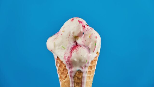 Timelapse of vanilla ice cream with strawberry topping and colorful sprinkles in waffle cone melting on blue background. Delicious white ice cream melting. Close-up of sweet dessert. Food concept