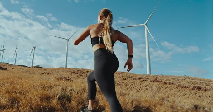 Slow motion of fitness woman running in field with windmills in the distance, green energy healthy sustainable future