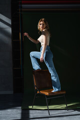 full length view of slim woman in jeans standing on chair on dark background.