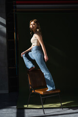full length of barefoot woman in jeans looking at camera while standing on chair on dark background.