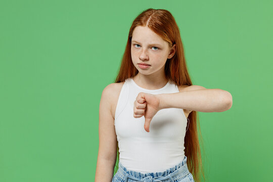 Little redhead kid disappointed sad girl 12-13 years old wearing white tank shirt showing thumb down dislike gesture isolated on plain green background studio portrait. Childhood lifestyle concept