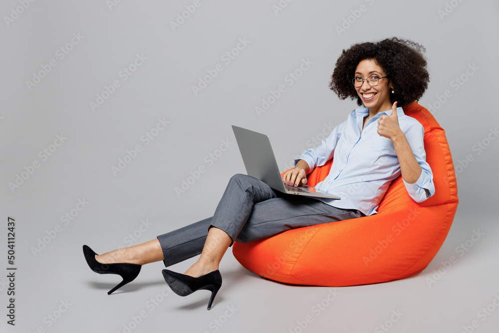 Wall mural Full body young employee business corporate lawyer woman of African American ethnicity in formal shirt work in office sit in bag chair hold use work on laptop pc computer isolated on grey background.