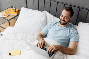 Top view young happy man wear blue t-shirt lying in bed hold use work on laptop pc computer rest relax spend time in bedroom lounge home in own room house wake up dream. Good mood bedtime concept