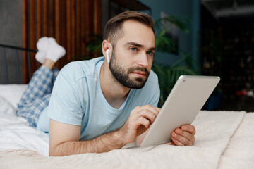 Full body pensive young man in blue t-shirt lying on bed listen music in earphones use tablet pc computer rest relax spend time in bedroom lounge home in own room house dream. Morning mood concept