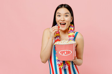 Excited amazed young woman of Asian ethnicity in striped one-piece swimsuit hawaii lei hold takeaway popcorn bucket isolated on plain pastel pink background. Summer vacation sea rest sun tan concept.