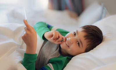 Obraz na płótnie Canvas Kid lying in bed using tablet reading story before bed time, Happy young boy playing game online or chatting with friends on internet, Child relaxing in bedroom in the morning