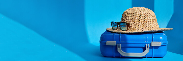 Plastic blue suitcase with straw hat and sunglasses placed on floor in bright studio for summer vacation banner