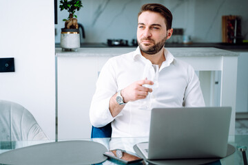 Businessman looking away, working on laptop in modern kitchen, checking email in morning. Drinking and relaxing at home