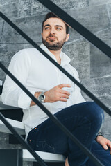 Vertical image attractive and masculine bearded man, sitting on stairs in loft. Holding glass vine in hands. Low angle