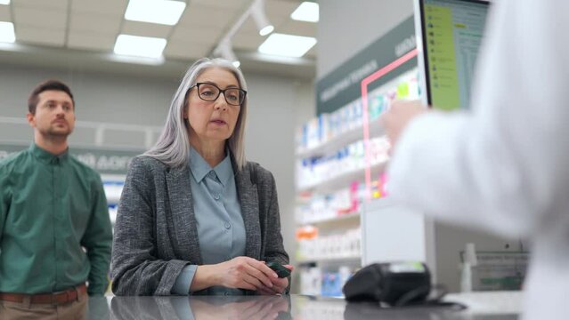 Senior caucasian woman with sore throat standing at modern apothecary and talking with qualified pharmacist while choosing pills. Concept of people, healthcare and pharmaceutics.