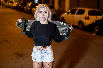 young pretty woman looking camera in shorts while standing on the street holding a skateboard at...