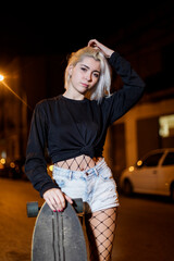 Front view of young pretty woman looking camera in shorts while standing on the street holding a skateboard at night in the city