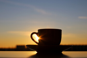 Black silhouette of coffee or tea cup on sunset sky background. View from the window to evening city