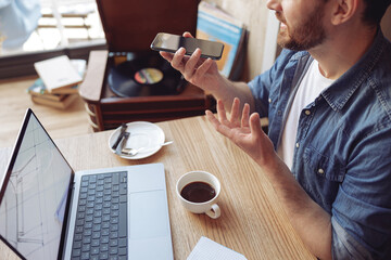 Mobile phone in male hand. Man chatting and sending voice message with laptop and coffee. Close up.