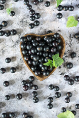 Freshly gathered black currants with leaves in a plate in shape of heart and on grey surface background inside, fresh black currants in dish, healthy eating concept, top view