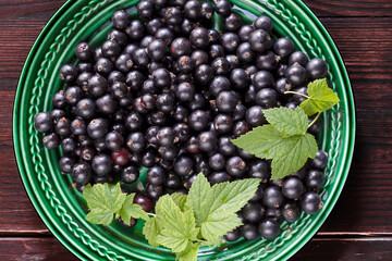 Freshly gathered black currants with leaves in a green plate and on wooden brown table background inside, fresh black currants in dish, harvest of black currants in rustic style, top view
