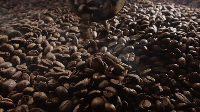 Coffee beans are roasted with little roasted smoke, the camera close up slowly follows the numerous coffee beans. Small amount of coffee beans spill out of the scoop. 