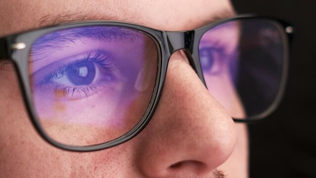Focused office worker wears black-rimmed glasses for reducing eye strain. Young man in spectacles looks at screen working on computer closeup