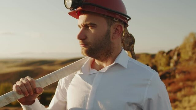 Businessman man in helmet with pickaxe on shoulder, looks to side and turns head to camera, side view. Businessman cryptocurrency miner stands outdoors in hard hat. Concept of cryptocurrency
