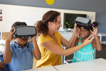 Caucasian young female teacher wearing vr glasses to african american elementary schoolgirl in class - Powered by Adobe