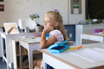 Caucasian elementary schoolgirl with hand on chin looking away while sitting at desk in classroom