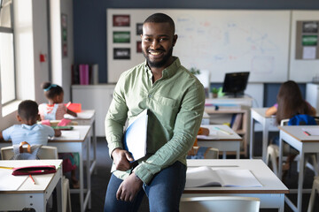 Portrait of smiling african american young male teacher holding file while sitting at desk in class - Powered by Adobe