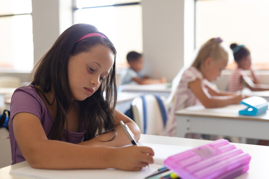 Caucasian elementary schoolgirl writing on book while studying at desk in classroom