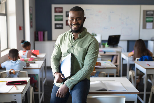 Portrait of smiling african american young male teacher holding file while sitting at desk in class