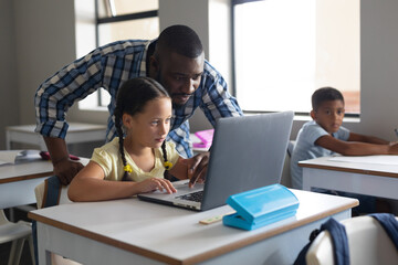 African american young male teacher showing laptop to caucasian elementary schoolgirl at desk