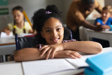 Portrait of smiling biracial elementary schoolgirl sitting on wheelchair at desk in classroom