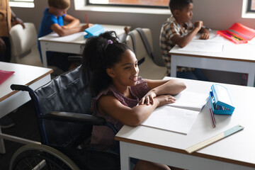 Smiling biracial elementary schoolgirl sitting on wheelchair at desk in classroom